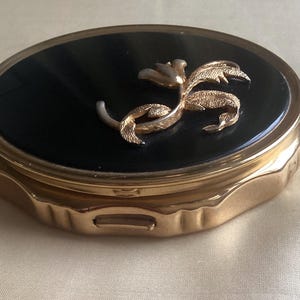 Könnte beinhalten: Ein ovaler, goldfarbener Kompaktspiegel mit schwarzem Deckel. Auf dem Deckel befindet sich ein goldfarbenes Blumendesign. Der Kompaktspiegel hat eine Klappöffnung und einen dekorativen, geriffelten Rand. Abmessungen in Zentimetern.