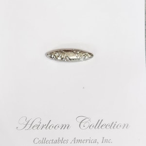 May include: A small, silver-toned brooch with an oval shape and ornate floral detailing. The brooch is centered on a white background, with the words "Heirloom Collection" and "Collectables America, Inc." printed below.