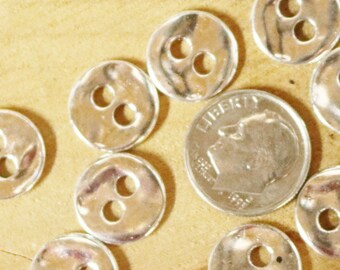 May include: A collection of silver-toned metal buttons with two holes each. The buttons are arranged in a circle on a wooden surface.
