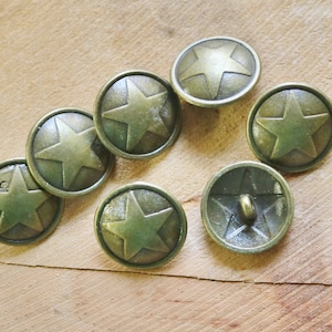 May include: Seven antique gold-toned metal buttons with a star design. The buttons are arranged in a semi-circle on a wooden surface.