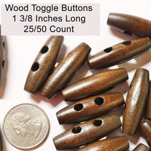 May include: A close-up of a pile of brown wooden toggle buttons. The buttons are 1 3/8 inches long and have two holes. The text "Wood Toggle Buttons 1 3/8 Inches Long 25/50 Count" is visible in the image.