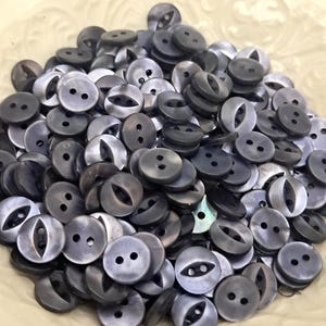 May include: A pile of dark gray buttons with two or four holes. Some buttons have an oval-shaped opening. The buttons have a pearlescent sheen and are round. The buttons are likely for sewing or crafts.