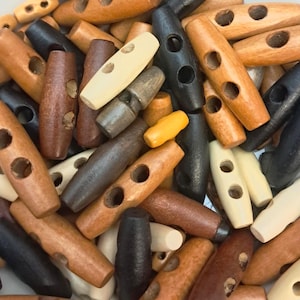May include: A collection of wooden toggle buttons in various shades of brown, black, and cream. The buttons are oblong with two holes for sewing and are made of natural wood.