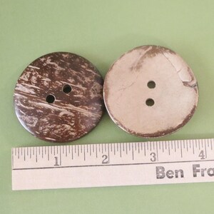 May include: Two large, round buttons made from coconut shell. One button is dark brown with a marbled pattern, and the other is light brown. Each button has two holes for sewing. A ruler is placed below the buttons.