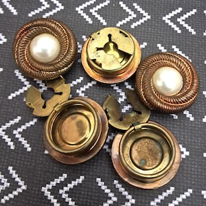 May include: An assortment of vintage brass and copper buttons. Two buttons feature a central pearl surrounded by a woven, copper-colored design. The other buttons have a circular design with a hinged clasp.