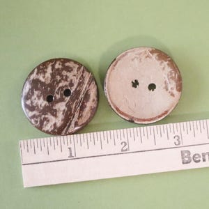 May include: Two large, round buttons made from a natural material, likely coconut shell, with a dark brown and cream marbled pattern. Each button has two holes for sewing. A ruler is placed next to the buttons.