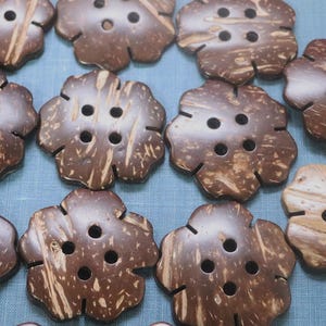 May include: A collection of flower-shaped buttons made from natural coconut shells. The buttons are various shades of brown with visible wood grain and four holes for sewing. They are arranged on a light blue surface.