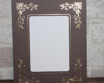 May include: A brown picture frame with a gold-colored floral design around the edges. The frame has a white mat and a clear opening for a photo.