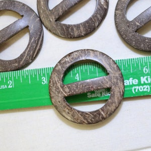 May include: Four brown coconut shell belt buckles with a center hole and a horizontal bar across the center. The buckles are arranged in a semi-circle on a white surface with a green ruler.