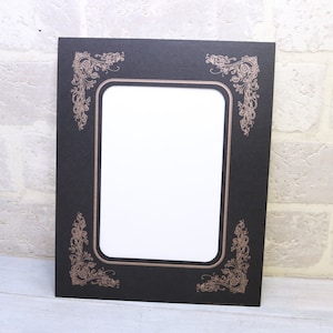May include: A black picture frame with a white mat and ornate gold-colored corners. The frame is empty.