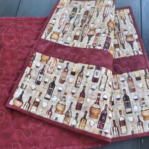 May include: A quilted table runner with a wine theme. The runner features a pattern of wine bottles and glasses on a beige background, framed by a burgundy border and backing. The burgundy fabric has a quilted design.