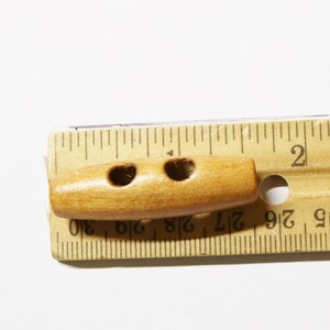 May include: A wooden toggle button with two holes, measuring approximately 1 inch in length. The button is light brown in color and is shown next to a ruler.