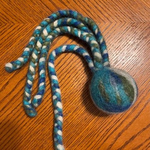 Puede incluir: Un juguete de lana fieltrada que presenta una bola redonda unida a cuatro tentáculos largos y trenzados. El objeto está elaborado con fibras mezcladas en tonos azul, verde azulado, verde y blanco, creando una apariencia jaspeada y multicolor en toda la esfera y los apéndices similares a cuerdas.