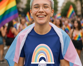 Op de afbeelding: Een persoon draagt een marineblauw t-shirt met een regenboogontwerp en het woord "PRIDE" onderaan. De persoon lacht en draagt een ronde bril. Een transgender vlag is over hun schouders gedrapeerd, met regenboogvlaggen op de achtergrond.