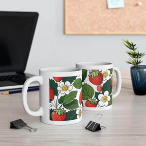 Pode incluir: Duas canecas de cerâmica branca com um padrão de morangos. O design apresenta morangos vermelhos, folhas verdes e flores brancas com centros amarelos. As canecas estão sobre uma superfície clara, com um laptop e material de escritório ao fundo.