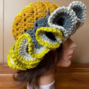 May include: A crochet hat featuring a mustard yellow crown with a textured stitch pattern. The wide, ruffled brim is layered with alternating bands of dark gray, light gray, and lime green yarn, creating a voluminous, wavy edge design.