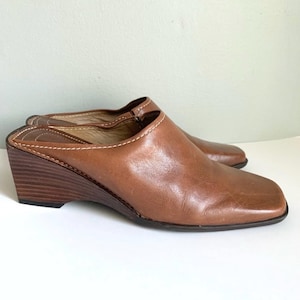 May include: Brown leather wedge heeled mules with a square toe. The shoes have a stitched seam around the top edge.