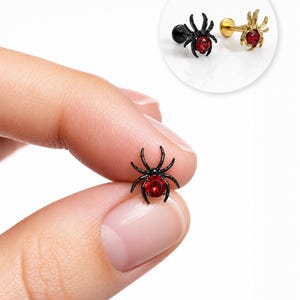 May include: A spider-shaped stud earring featuring a black metal body and a round, faceted red gemstone at its center. The design includes eight thin, curved legs extending from the central stone. A secondary gold-toned version is also shown.