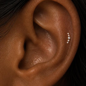 May include: A gold-toned curved cartilage earring featuring five small, clear, round-cut stones arranged in a graduated size pattern. The jewelry is positioned along the upper ridge of the ear, following the natural curve of the cartilage.