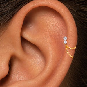 May include: A gold ear piercing featuring two small, clear, round-cut stones set vertically, with two delicate gold chains draping from the lower stone to create a looped effect. The text ORLA STUDIO is visible in the bottom right corner.