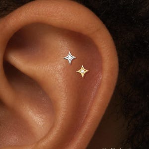 Op de afbeelding: Twee stervormige oorpiercings zijn geplaatst op het bovenste kraakbeen van het oor. De ene knop is zilverkleurig en de andere goudkleurig, elk met een kleine, heldere, centrale edelsteen. De tekst ORLA STUDIO is zichtbaar in de rechterbenedenhoek.