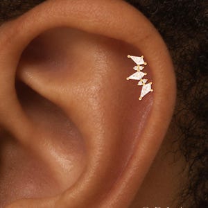 May include: A gold-toned ear cartilage stud featuring three kite-shaped clear stones arranged in a vertical cluster, interspersed with two small round clear stones. The jewellery is positioned on the upper ear cartilage. The text ORLA STUDIO is visible.