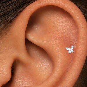 May include: A silver-toned butterfly-shaped ear stud featuring four clear, faceted stones arranged to form wings. The jewelry is positioned on the cartilage of an ear. The text ORLA STUDIO is visible in the bottom right corner of the image.