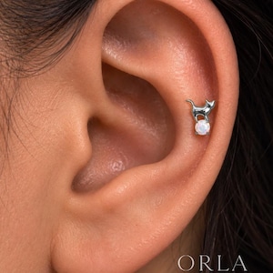 May include: A silver-toned cat-shaped earring stud featuring a small, round, iridescent white opal stone positioned beneath the cat's body. The jewelry piece is secured in the upper cartilage area of an ear, displaying a polished metallic finish.