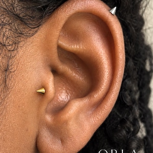 May include: Two cone-shaped stud earrings are worn in the ear. One earring is gold-toned and positioned in the tragus, while the other is silver-toned and located on the upper cartilage. The text ORLA STUDIO is visible in the bottom right corner.