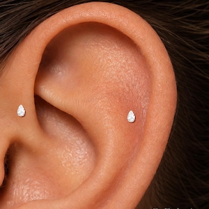 May include: Two small, silver-toned stud earrings, each featuring a clear, pear-shaped gemstone. The earrings are positioned in the ear, one in the tragus and one in the conch area. The text ORLA STUDIO is visible in the bottom right corner.