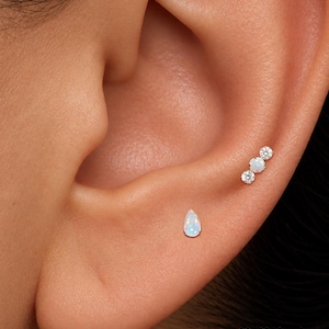 May include: Two silver-toned ear piercings are visible. One is a teardrop-shaped opal stud, and the other is a linear cluster featuring a central opal flanked by two clear round stones. The text ORLA STUDIO is printed in the bottom right corner.