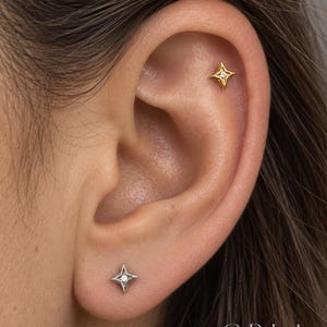 May include: A gold star-shaped stud earring with a central clear stone in the upper ear cartilage and a silver star-shaped stud earring with a central clear stone in the earlobe. The text ORLA STUDIO is visible in the bottom right corner.