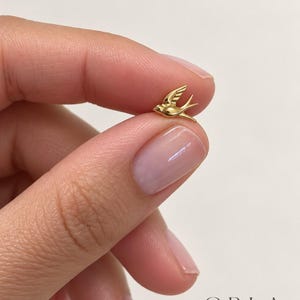 May include: A small, polished gold-tone swallow bird stud earring featuring a detailed wing design and a pointed tail. The piece is crafted with a smooth, reflective surface and is positioned to show its profile, with the text ORLA STUDIO visible.