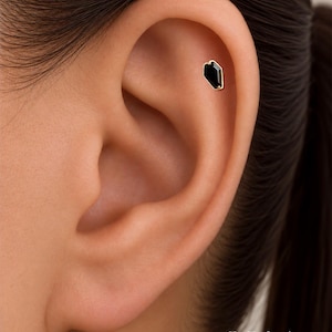 May include: A gold stud earring featuring a black, coffin-shaped gemstone set in a metal frame. The earring is positioned in the upper cartilage area of an ear. The text ORLA STUDIO is visible in the bottom right corner of the image.