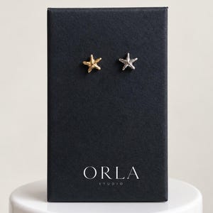 May include: A pair of stud earrings shaped like starfish, one in a gold tone and one in a silver tone, displayed on a rectangular black card with the white text ORLA STUDIO printed at the bottom center of the card surface.