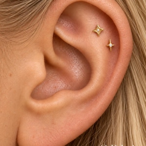 May include: Two gold-toned ear studs in the shape of four-pointed stars, each featuring a small, clear, central gemstone. The studs are positioned in the upper cartilage area of an ear, with the text ORLA STUDIO visible in the bottom corner.