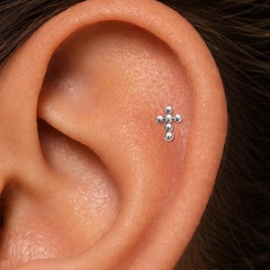 May include: A silver-toned stud earring shaped like a cross, constructed from five small, polished spherical beads arranged in a vertical and horizontal pattern, positioned securely in the upper cartilage of an ear.