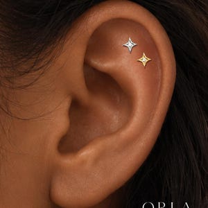 Könnte beinhalten: Zwei sternförmige Ohrstecker befinden sich im oberen Knorpelbereich des Ohrs. Ein Ohrstecker ist silberfarben mit einem kleinen Stein in der Mitte, der andere ist goldfarben mit einem kleinen Stein. Der Text ORLA STUDIO ist unten zu sehen.