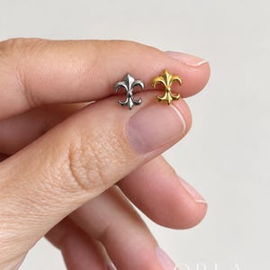 May include: Two small stud earrings shaped like fleur-de-lis symbols. One earring is finished in a polished silver tone, while the other is finished in a polished gold tone. Both pieces feature a classic three-petal design with a central vertical element.