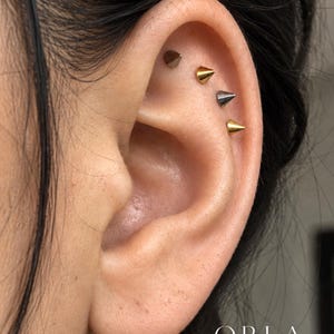 May include: Four small, cone-shaped stud earrings arranged in a vertical row along the upper cartilage of an ear. The studs alternate between metallic gold and dark gunmetal gray finishes. The text ORLA STUDIO is visible in the bottom right corner.