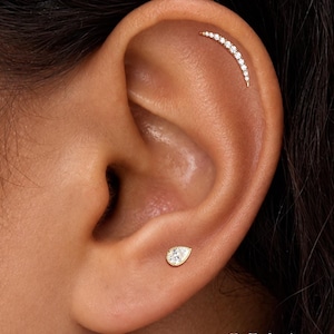 May include: A gold ear piercing set featuring a curved bar stud with small clear stones along the upper cartilage and a single pear-shaped stud with a clear stone in the lobe. The text ORLA STUDIO is visible in the bottom right corner.