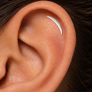 May include: A silver crescent moon shaped ear piercing jewelry piece positioned along the upper cartilage of an ear. The jewelry has a smooth, metallic finish and a curved design. The text ORLA STUDIO is visible in the bottom right corner.