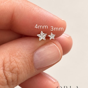 Puede incluir: Dos pendientes de botón en forma de estrella plateada con piedras transparentes. Un pendiente está etiquetado como 4mm y el otro como 3mm. Los pendientes son de plata metálica con piedras transparentes facetadas. El texto ORLA STUDIO es visible.
