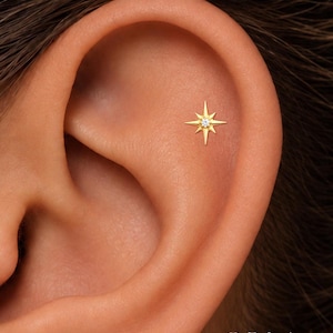 May include: A gold-toned ear piercing stud featuring a starburst design with a central clear gemstone. The jewelry is positioned in the upper cartilage area of an ear. The text ORLA STUDIO is visible in the bottom right corner of the image.