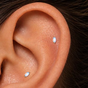 Könnte beinhalten: Zwei marquiseförmige Ohrringe mit irisierenden, mondsteinartigen Edelsteinen in goldfarbenem Metall. Ein Ohrring befindet sich im Helix, der andere im Tragus. Der Text ORLA STUDIO ist unten rechts im Bild zu sehen.