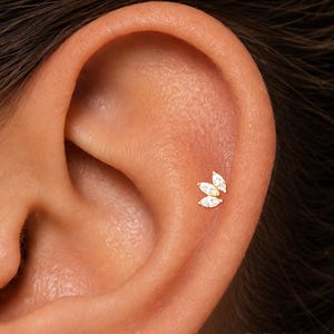 May include: A gold-toned ear stud featuring three marquise-cut clear stones arranged in a cluster. The jewelry is positioned on the cartilage of an ear. The text ORLA STUDIO is visible in the bottom right corner of the image.