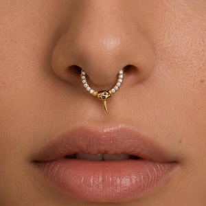 Puede incluir: Un anillo de septum dorado con una banda curva incrustada de pequeñas piedras transparentes. El centro del anillo muestra un diseño de cabeza de serpiente con dos diminutos ojos negros y una cola puntiaguda orientada hacia abajo.