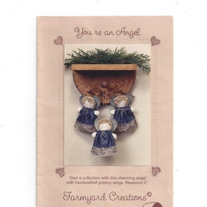 May include: A handmade angel ornament with blue fabric wings and a white lace trim. The angel is hanging from a wooden shelf with a green plant. The text "You're an Angel" and "Farmyard Creations" is printed on the page. The angel measures 5 inches.
