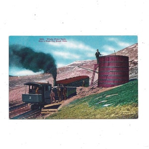 May include: A vintage postcard depicting a train on the Pike's Peak Cog Railway in Colorado. The train is a small, steam-powered locomotive with a water tank on top. The train is traveling up a steep, rocky incline. The postcard is titled "633. Windy Point Tank, Pike's Peak Cog Road, Colo."