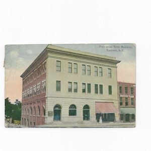 May include: Vintage postcard featuring the Enterprise Bank Building in Laurens, S.C. The building has a cream-colored facade with multiple windows and a red awning. The postcard is slightly aged, with a faded appearance.